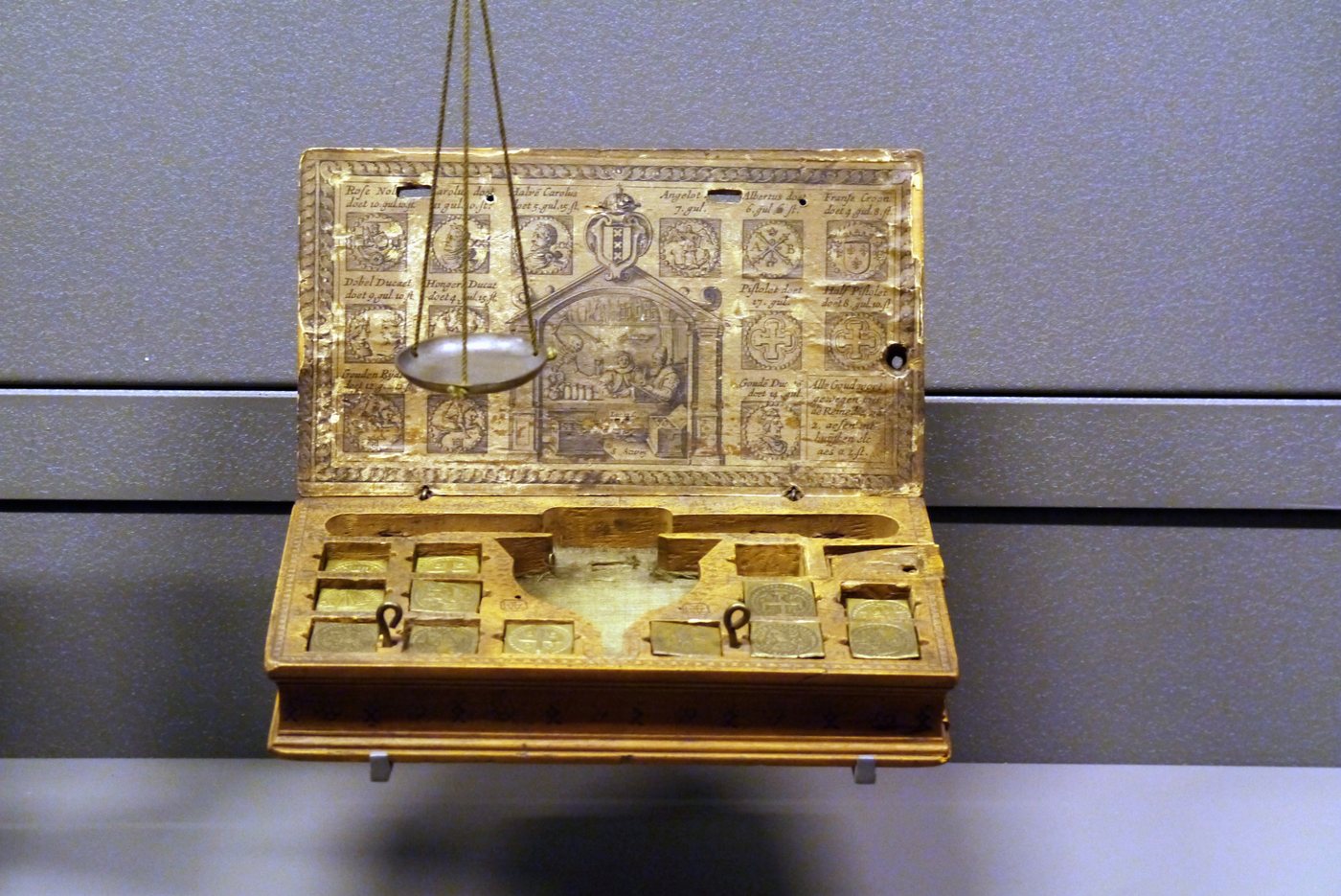 A small wooden box holding a balance scale and a set of nine brass weights.