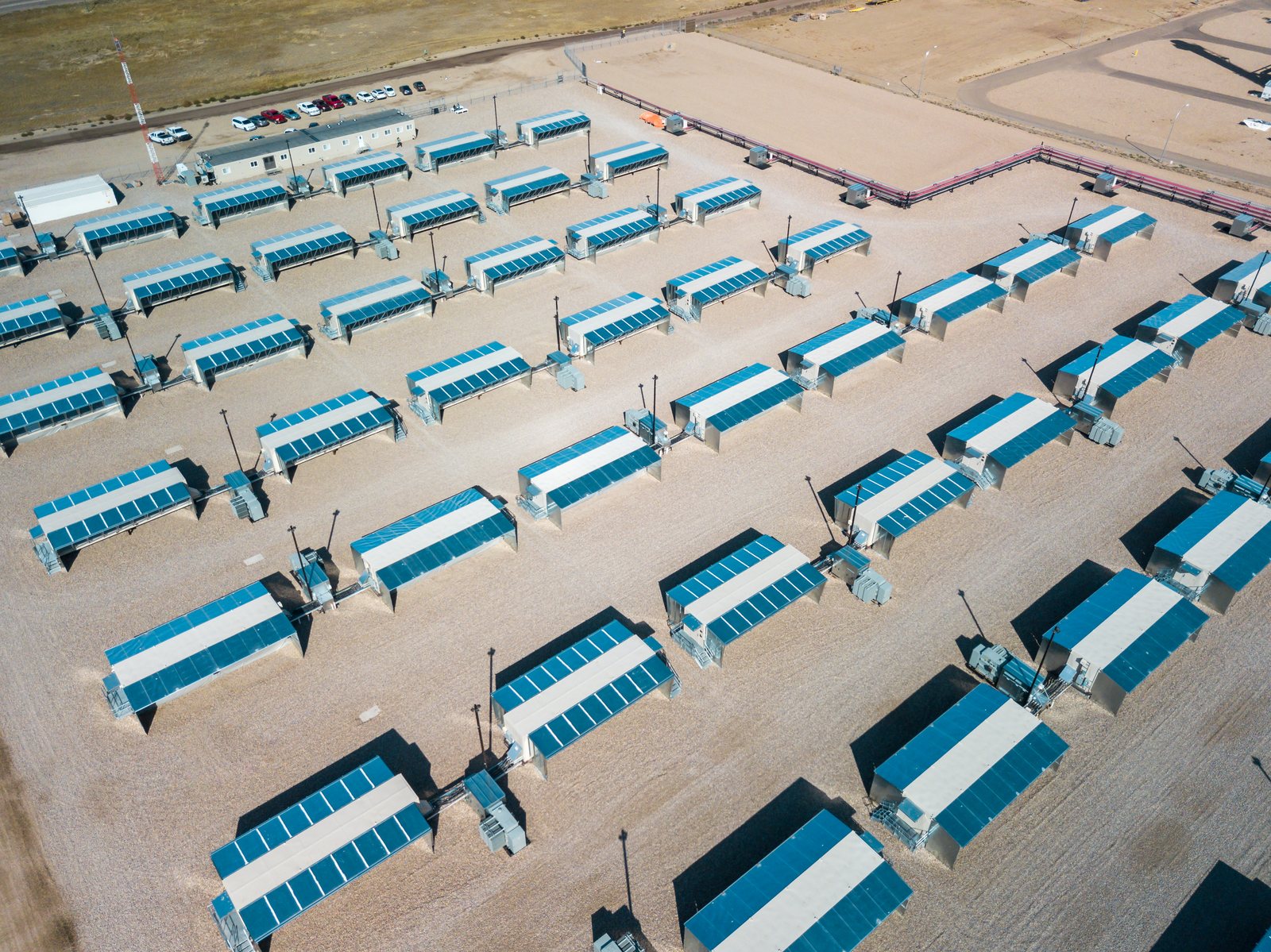 Aerial view of a large industrial bitcoin mining facility, rows of long buildings and containers on a bare site.