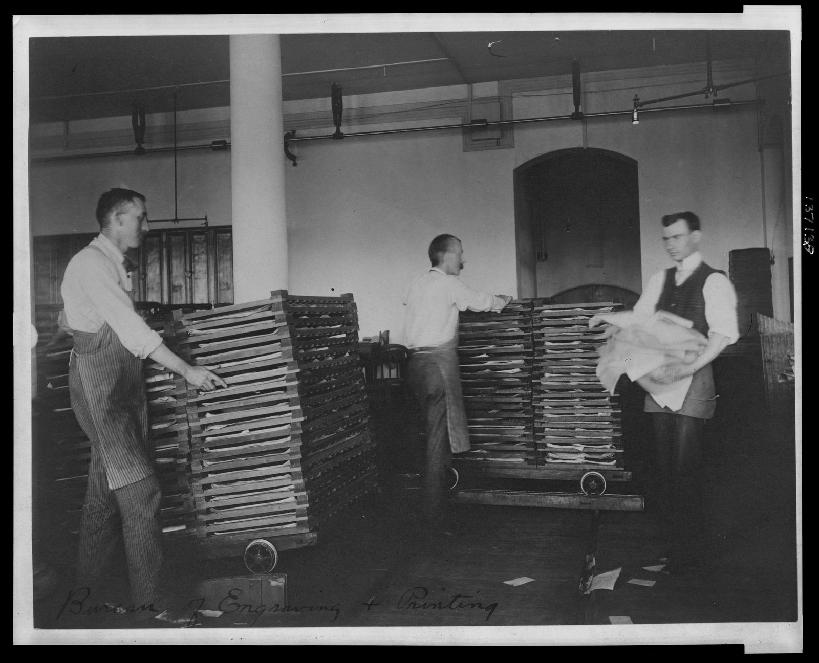 Two workers in a nineteenth-century workshop carrying large sheets of freshly printed banknotes.