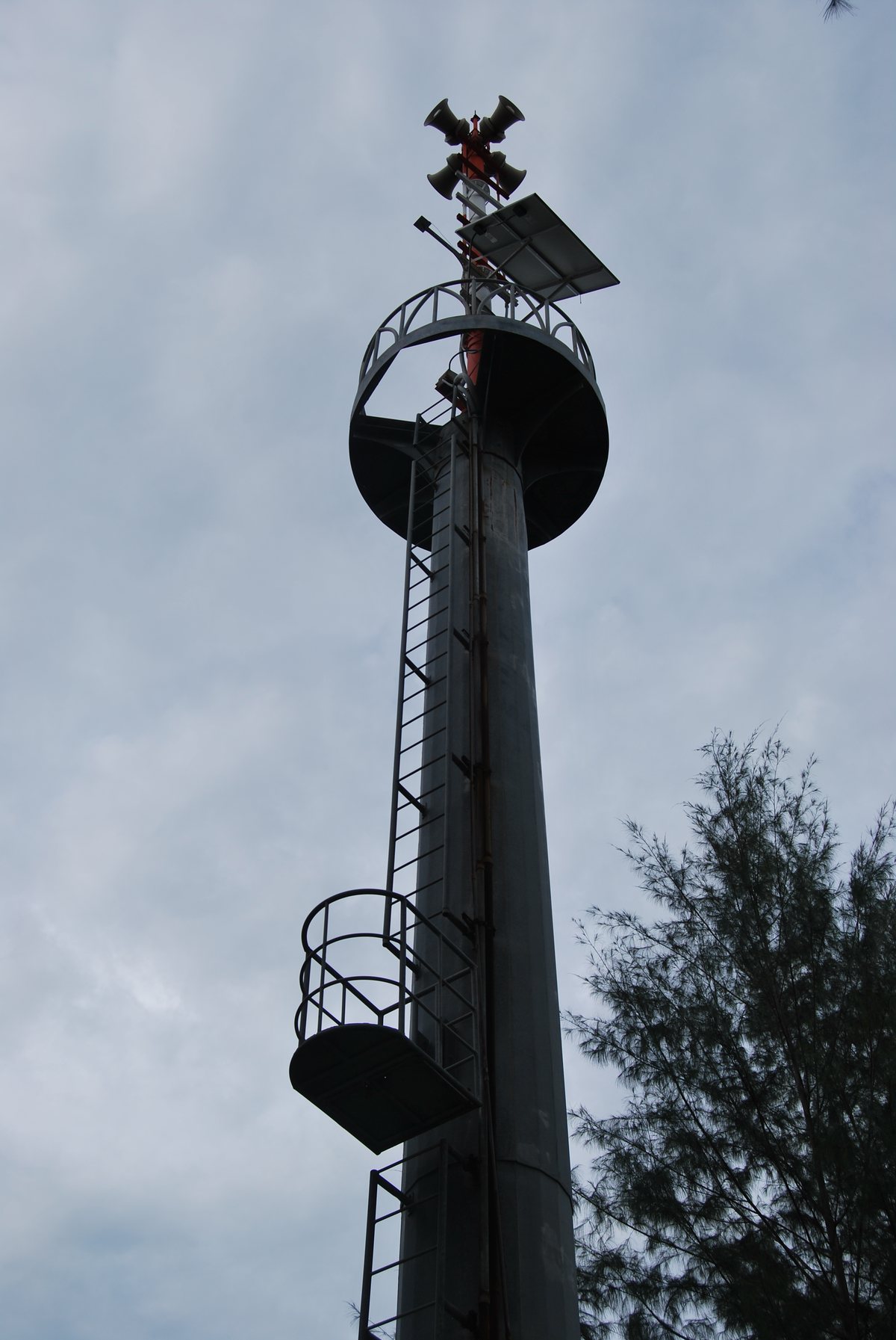 A tall metal tsunami warning tower with loudspeakers, standing above palm trees near Khao Lak