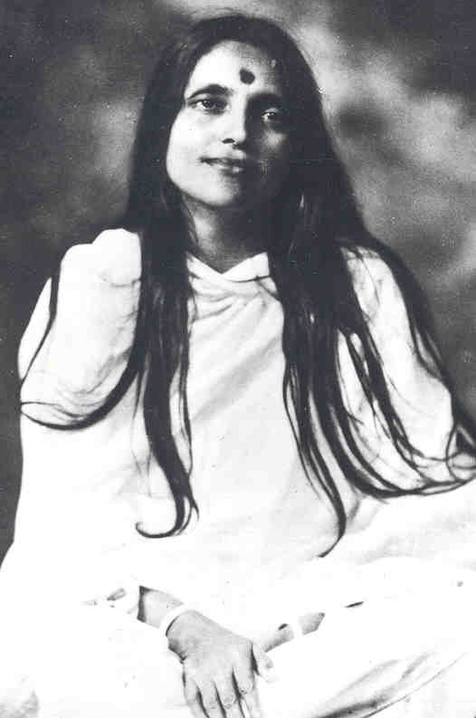 A black-and-white photograph of the young Anandamayi Ma, seated, with long dark hair and a calm direct gaze.