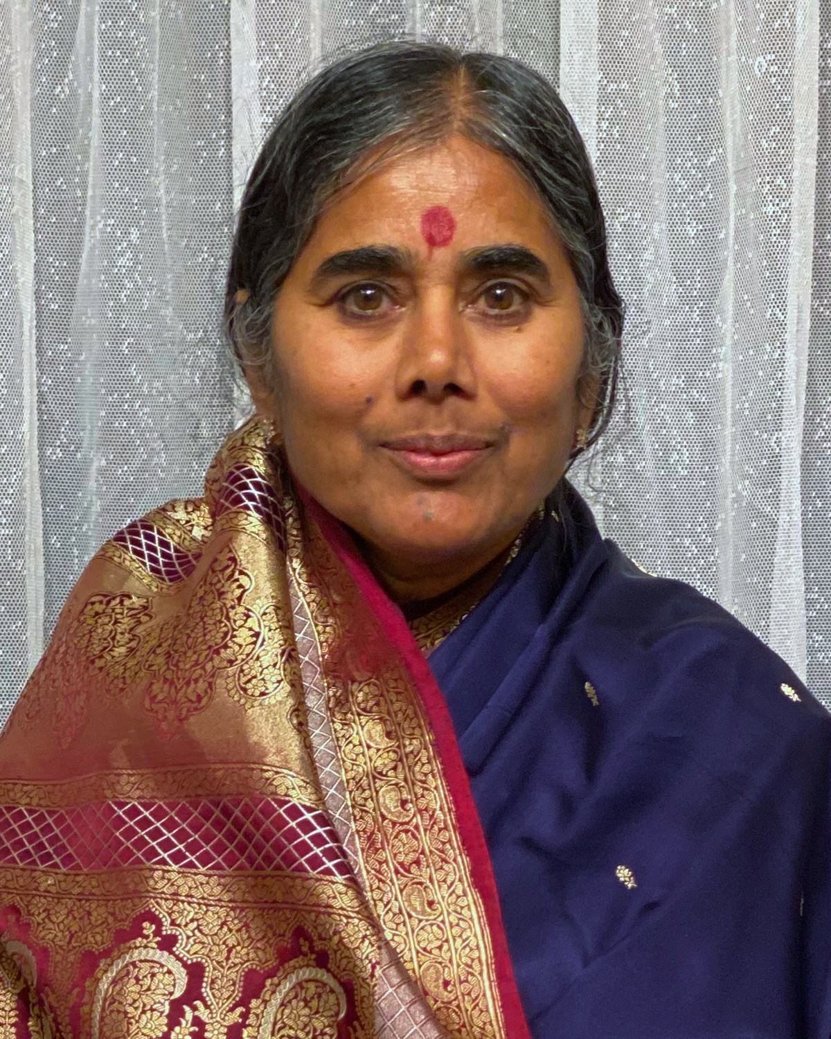 A colour photograph of Mother Meera in 2020, wearing a dark blue sari with a red and gold shawl, looking calmly at the camera.