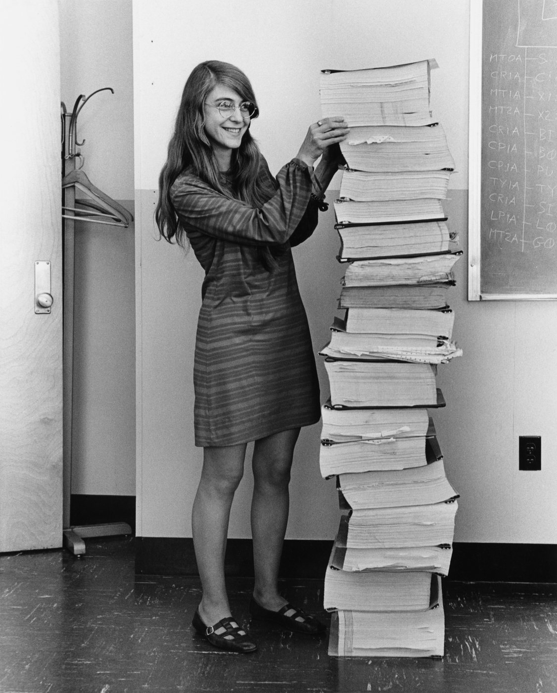 Margaret Hamilton standing beside a stack of printed Apollo program listings as tall as she is
