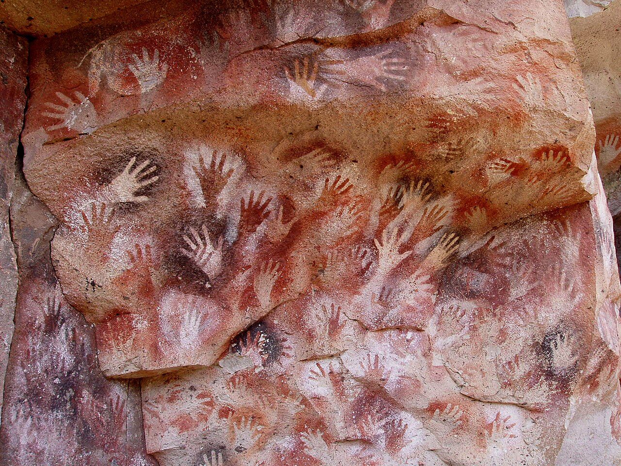 Dense overlapping stencilled handprints in red, ochre and white on a rock wall at Cueva de las Manos, Argentina.