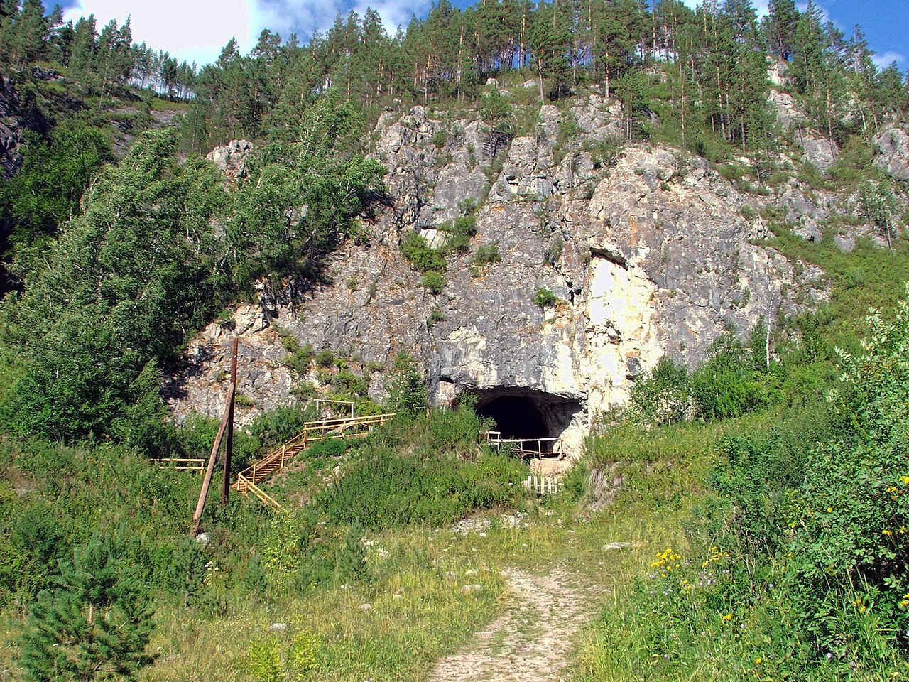 The entrance to Denisova Cave in the Altai Mountains, a dark opening in a limestone bluff surrounded by pine forest.