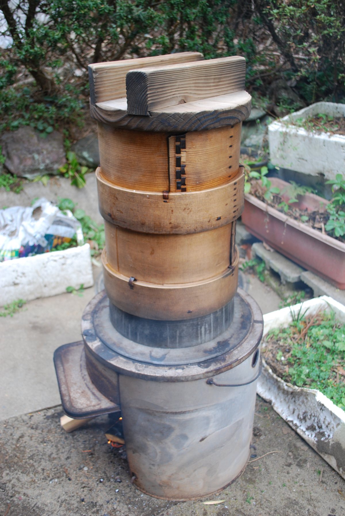A stack of cedar steaming baskets sitting over a small wood-fired stove, with the fire visible underneath.