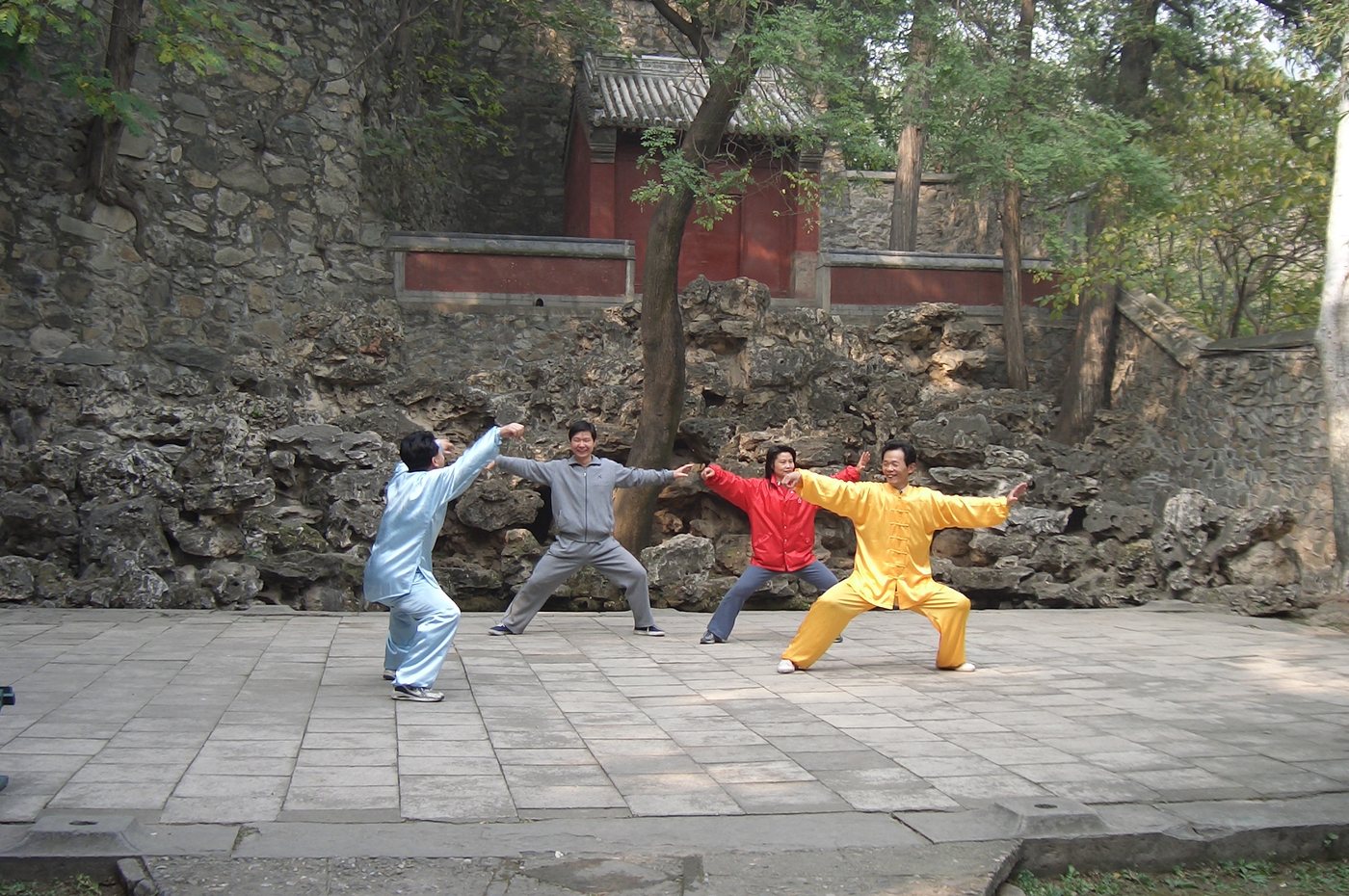 Four adults in a stone-walled park courtyard, arms extended in a wide low stance, practising Chen-style taijiquan together.